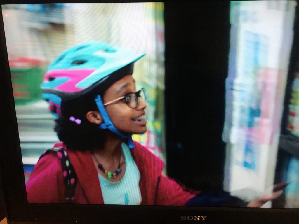 Thandi Ines, in a bike helmet, glasses, and red jacket, speaks in a scene from High Maintenance photographed off a TV screen