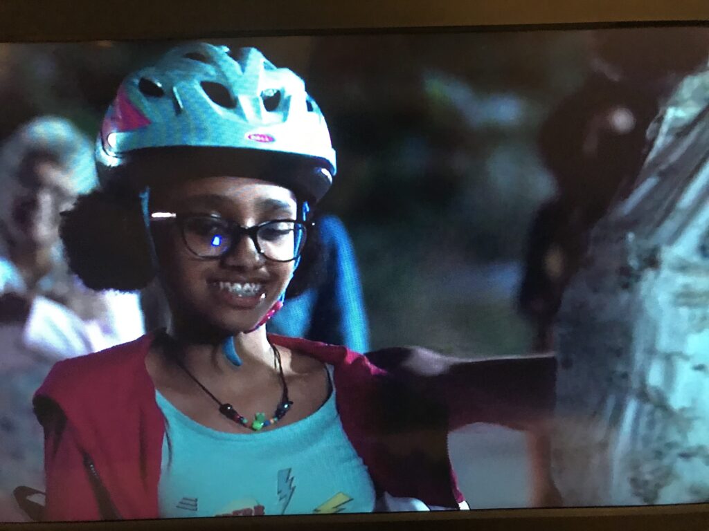 Thandi Ines grins in a blue bike helmet and glasses at night, in a scene from High Maintenance photographed off a TV screen