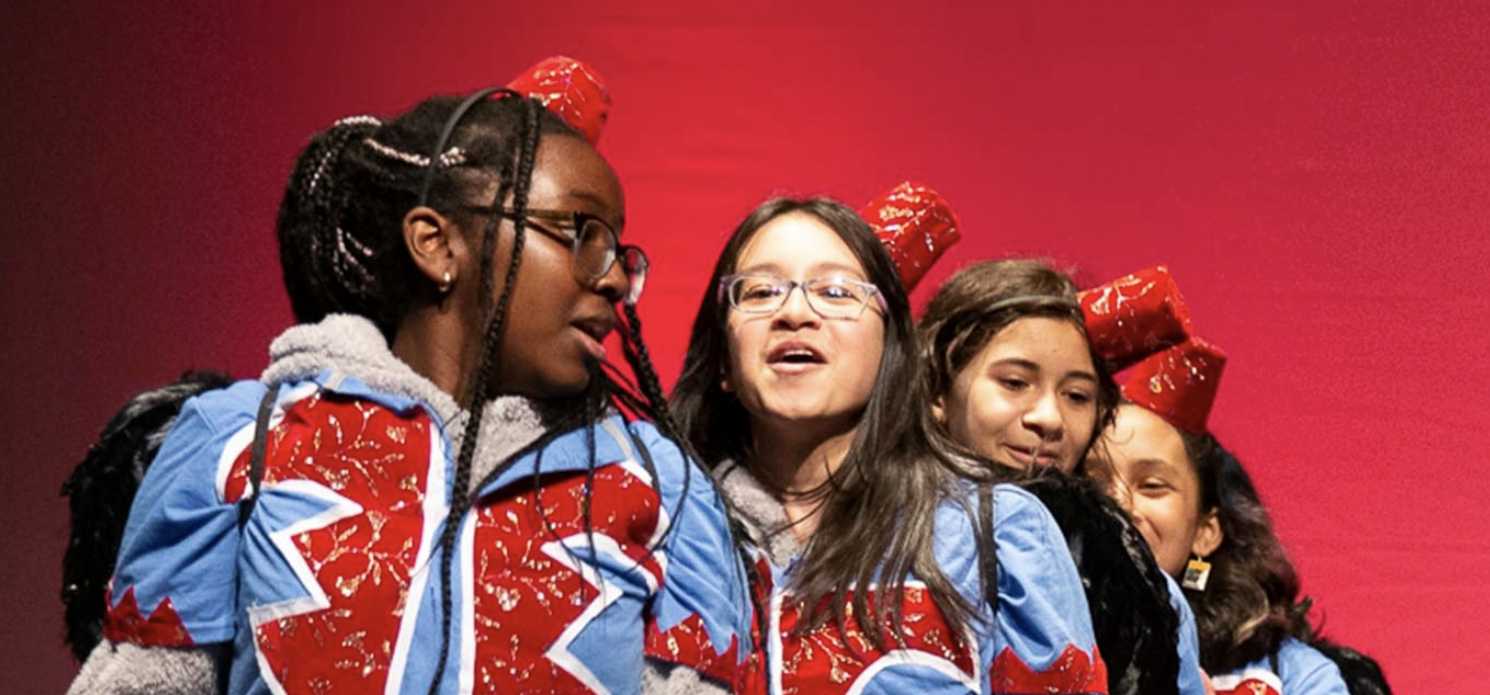 Four students in matching red and blue jackets and red caps lean in close and sing on stage against a red backdrop