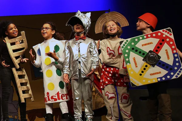 Young performers on stage in homemade costumes based on board games, including a Twister mat and a ladder