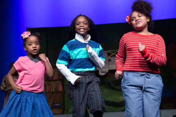 Three young performers step forward and sing on a stage lit in blue