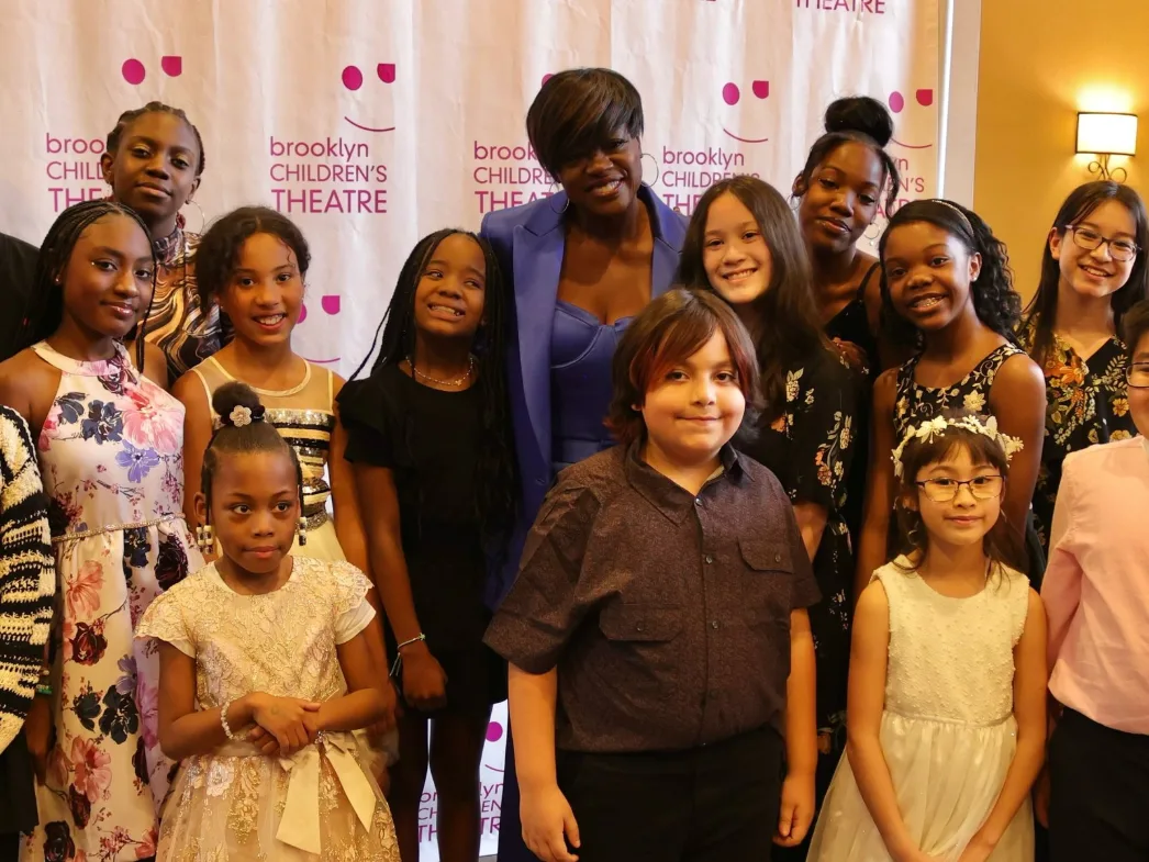 Viola Davis smiles in the middle of a group of about twenty children and teens in front of the Brooklyn Children’s Theatre backdrop