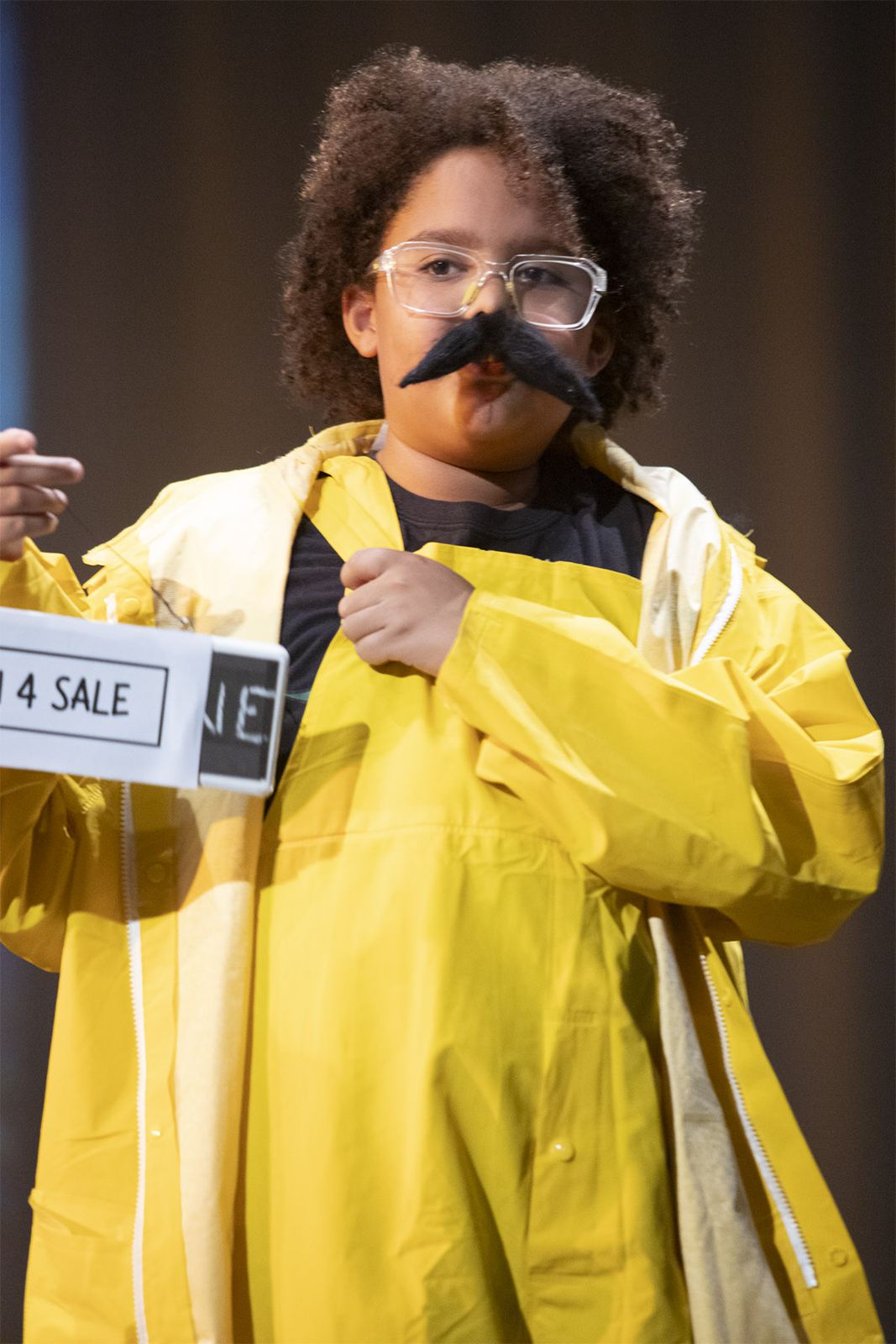 A student in a yellow raincoat and a fake mustache holds up a sale sign in Sirenia, Spring 2026