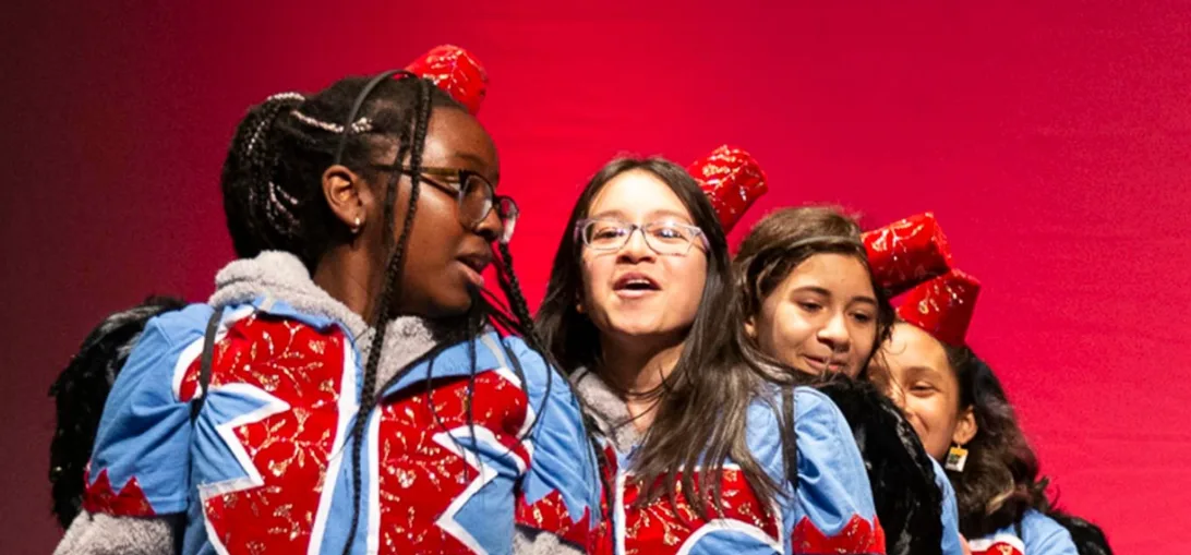 Four students in matching red and blue jackets and red caps lean in close and sing on stage against a red backdrop
